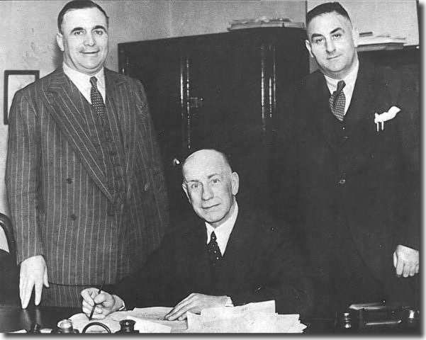 Chairman Sam Bolton and director Harold Marjason stand over Major Frank Buckley as he signs his contract as Leeds United manager